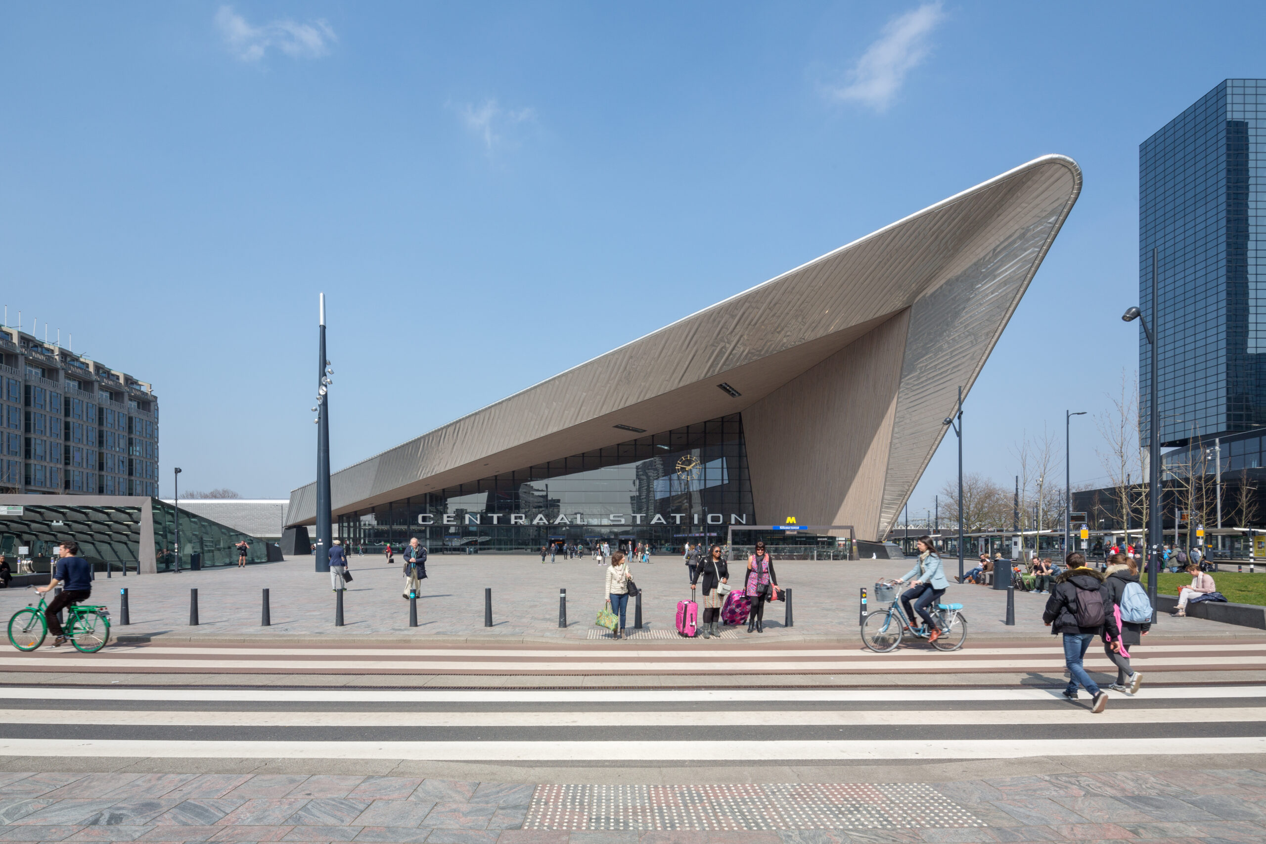 Voorplein van Rotterdam Centraal Station op een zonnige dag, met reizigers, fietsers en wandelaars voor de hoofdingang, foto: Ossip van Duivenbode